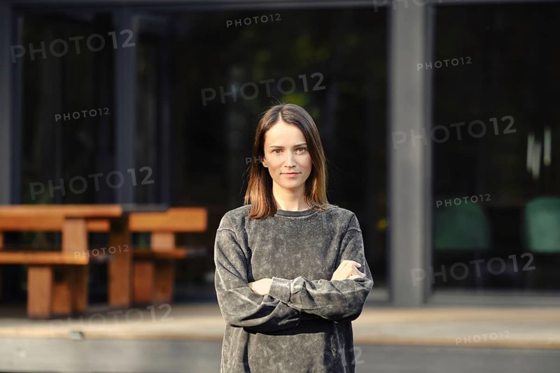A young woman with shoulder-length hair stands confidently with her arms crossed outside a sleek, modern building