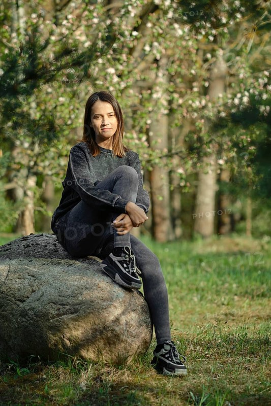 A young woman with long hair sits thoughtfully on a large rock in a peaceful forest. She wears casual clothing, enjoying the tranquility of nature. Sunlight filters through the trees