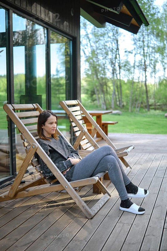 A woman sits comfortably in a wooden deck chair, enjoying the tranquil atmosphere. The lush green landscape and bright sunlight create a serene environment, perfect for relaxation