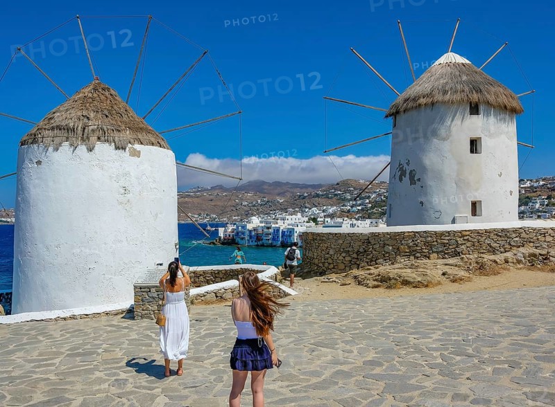 Mykonos, Cyclades, Greece - Tourists photograph the six sixteenth-century windmills lined up on a hill above Mykonos Town, Mykonos Chora. The windmills are the island's landmark. At the back, right by the sea, are the colorful houses of Little Venice. Mykonos is part of the Cyclades archipelago in the Aegean Sea