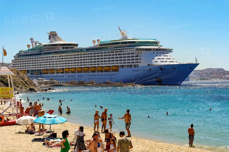 Mykonos, Cyclades, Greece - Voyager of the Seas cruise ship is located in Mykonos Harbour Bay. In front, tourists and locals bathe on the sandy beach of Paralia Agios Stefanos