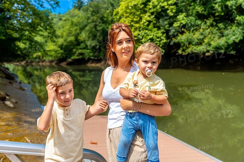 Mother holding her baby while walking alongside her other son on a picturesque pier, enjoying a sunny day by the river, surrounded by vibrant greenery and the beauty of nature