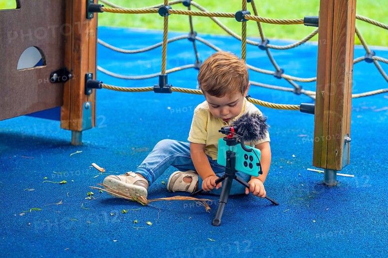 Little child sitting on playground's rubberized floor setting up a smartphone on a tripod with a microphone attached, creating videos and digital content