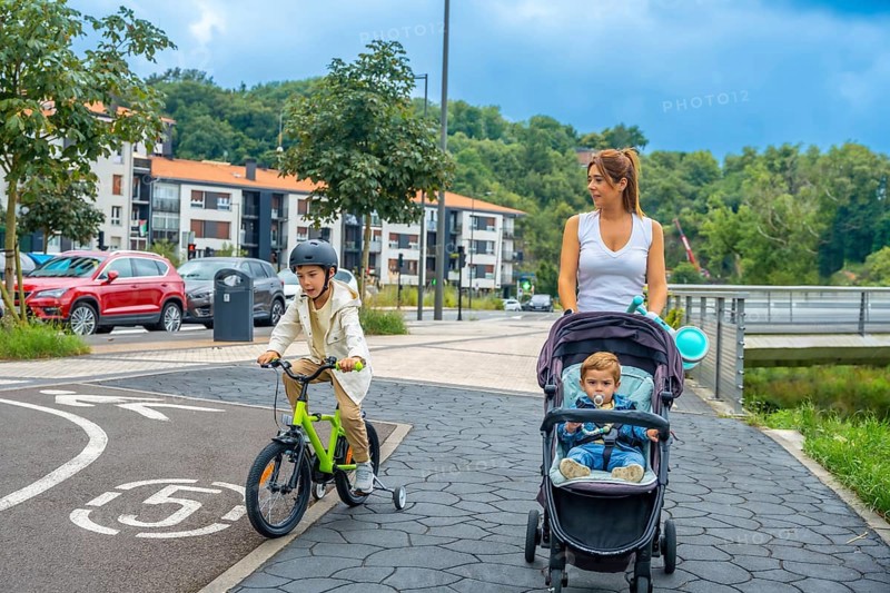 Mother pushing a baby stroller while her son rides a bicycle on a bike path in a modern urban environment, promoting healthy lifestyle and family bonding