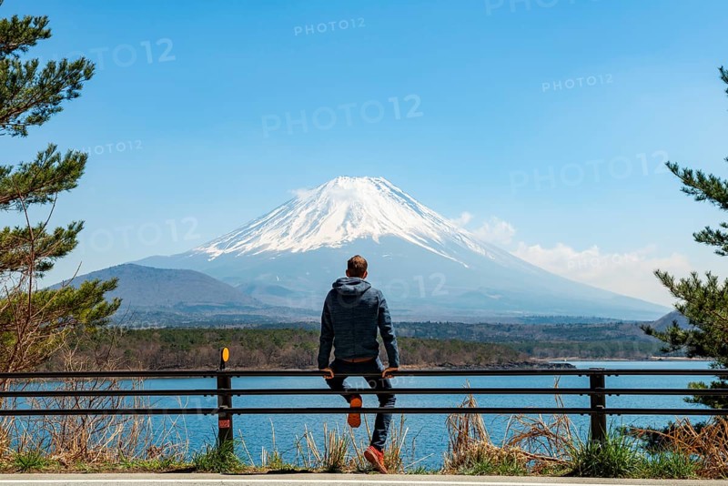 Young man sitting on railings next to a road and looking across the lake to Mt Fuji volcano, Motosu Lake, Yamanashi Prefecture, Japan