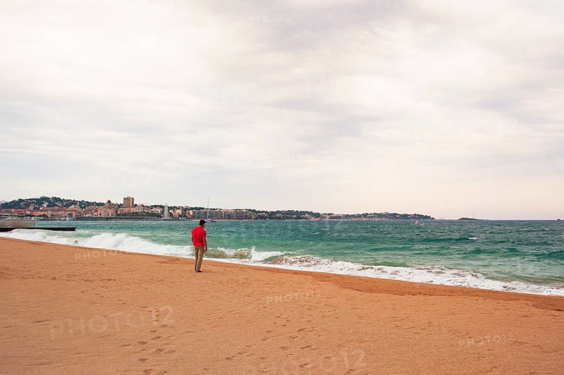Man wearing a red shirt looking at the moving sea, Fréjus in the background, Provence-Alpes-Côte d'Azur region, France
