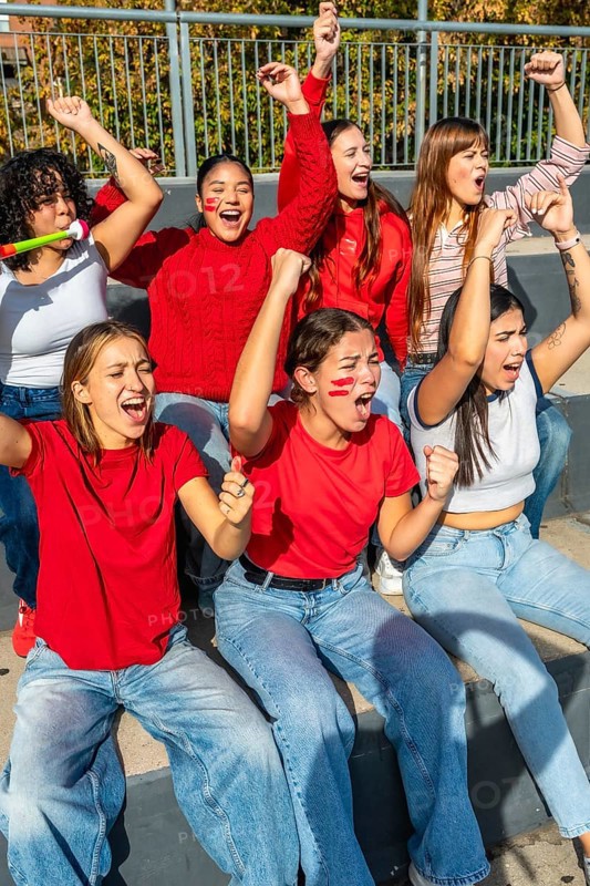 Young women sitting on stadium bleachers, cheering loudly with raised arms and painted faces, expressing excitement and support for their favorite team during a match