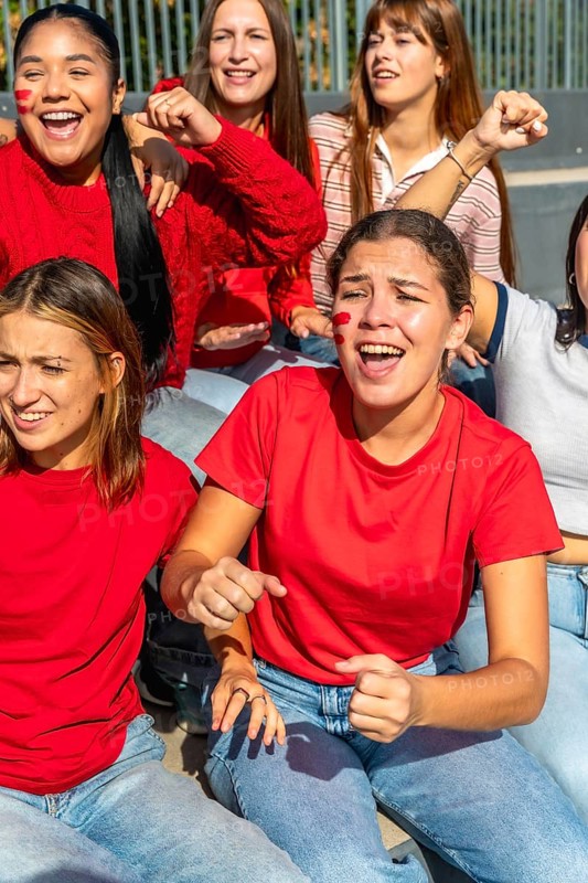 Group of enthusiastic young women friends wearing red shirts and face paint, actively cheering and expressing excitement while sitting together at a sports stadium