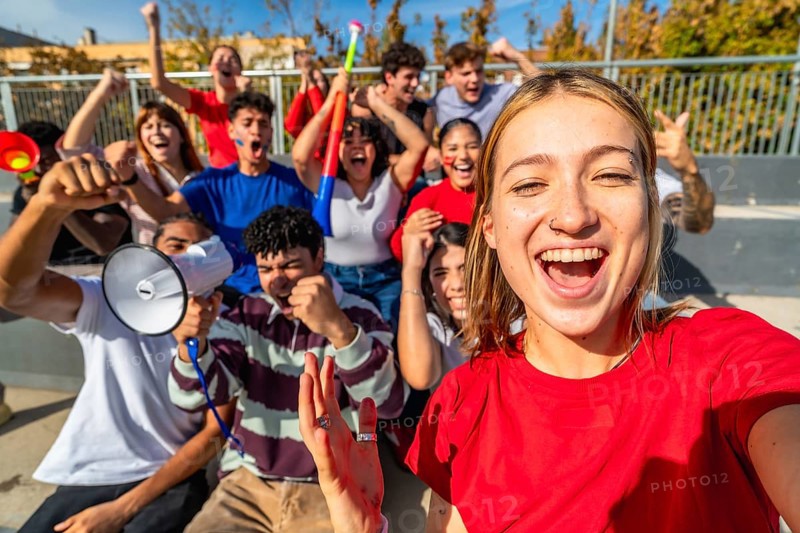 Young diverse fans cheering and taking a selfie in sunny stadium stands, celebrating their team with joyful smiles, energy and friendship during an exciting match