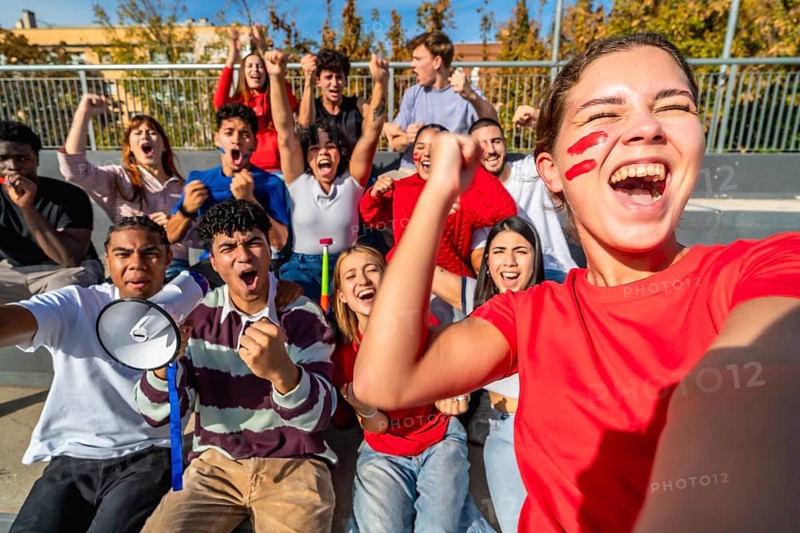 Diverse group of young adult fans energetically cheering and celebrating a win at a sports event, showing excitement, team spirit, and togetherness while watching from stadium bleachers