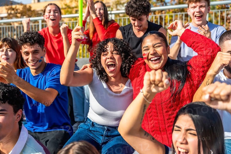 Diverse group of enthusiastic young people celebrating their team's victory at a stadium, waving hands, cheering loudly, and displaying team spirit with painted faces