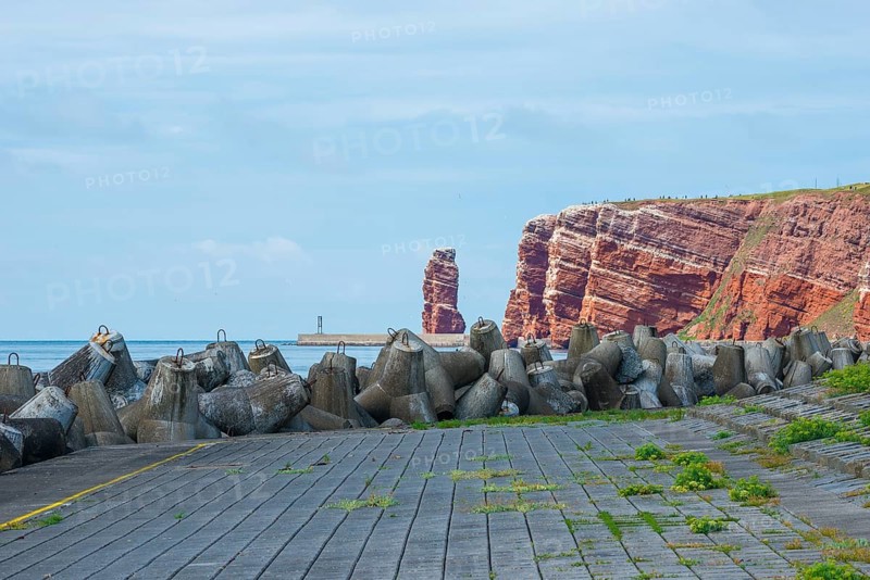 Sunny coastline with concrete tetrapods by the sea, coastal protection, protective wall, paved coastal strip with yellow line, danger, marking, warning, red cliffs and Lange Anna in the background, coastal path on the cliff, many people on the cliffs, rock layers in red sandstone, surf pillar, escape line, perspective, cloudy sky, blue sea on the horizon, western cliffs Poppe, high-seas island of Heligoland, North Sea, Pinneberg District, Schleswig-Holstein, Germany