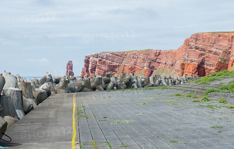 Coastline with concrete tetrapods by the sea, coastal protection, protective wall, paved coastal strip with yellow line, danger, marking, warning, red cliffs and Lange Anna in the background, cliff, cliffs, rock layers in red sandstone, surf pillar, escape line, perspective, overcast sky, blue sea on the horizon, west cliff, high-seas island of Heligoland, North See, Pinneberg District, Schleswig-Holstein, Germany