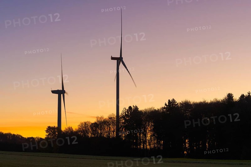 Two wind turbines in front of an orange-purple sky. Trees form a dense silhouette at dusk, Swabian Jura, Baden-Württemberg, Germany