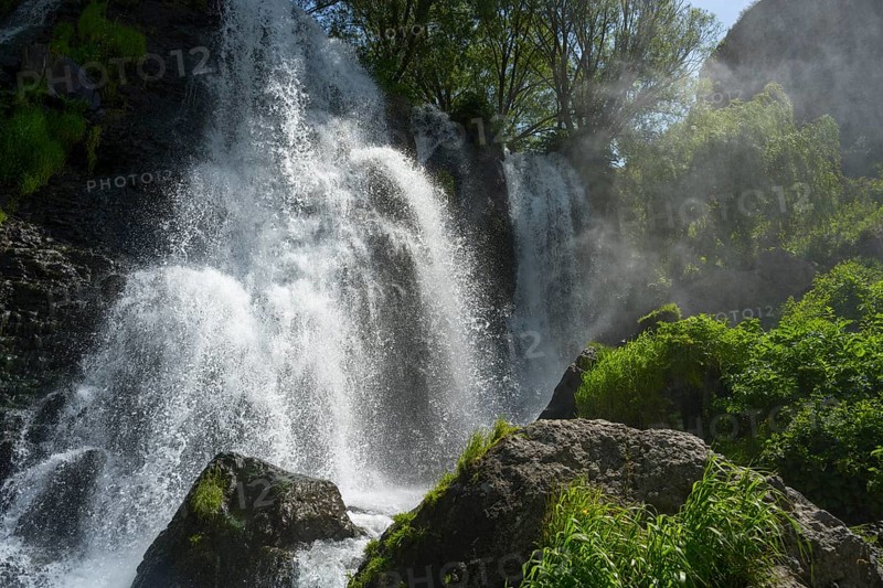 An impressive waterfall falls over rocks surrounded by green vegetation and trees in sunlight, Shaki Waterfall, Syunik Province, Syunik, Armenia