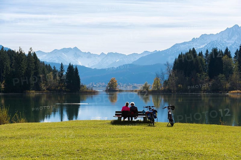 Two woman on a bench at Lake Forggensee enjoy the view of the mountains and the autumn landscape, bicycles are standing by Roßhaupten, Ostallgäu, Allgäu, Bavaria, Germany