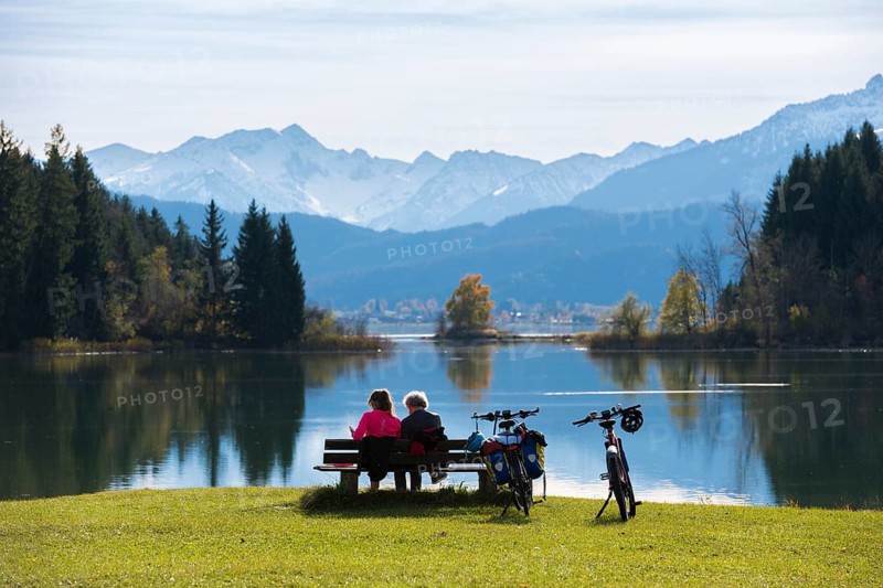 People sitting on a bench with a view over Lake Forggensee to Bergen, surrounded by peaceful nature and bicycles, near Roßhaupten, Ostallgäu, Allgäu, Bavaria, Germany