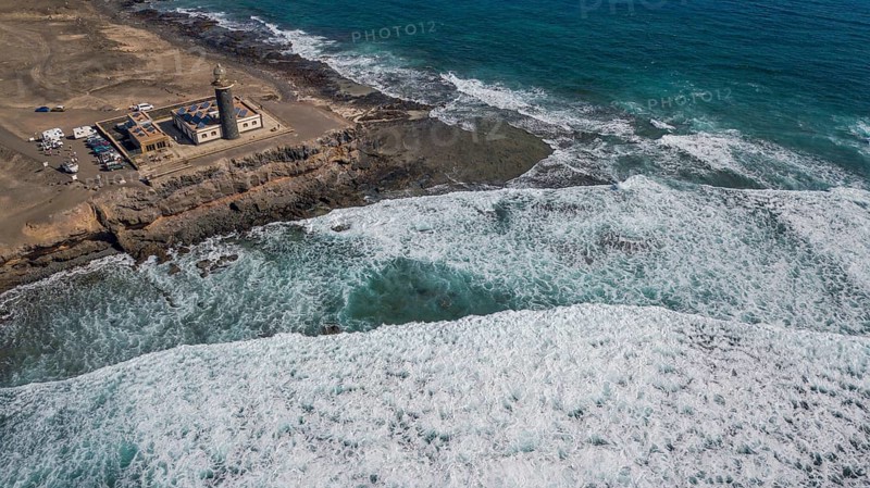 Aerial view of Faro Punta de Jandia lighthouse at the southern tip of Jandia peninsula, in the foreground white foam crowns on moving sea swell large high waves on reef edge beaming sea strong surf, Fuerteventura, Canary Islands, Canary Islands, Spain