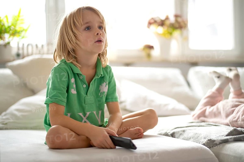 Child, boy, 7 years old, blonde, sitting on sofa, with remote control in hand, watching TV, watching television, watching television, excited, fascinated, Stuttgart, Baden-Württemberg, Germany