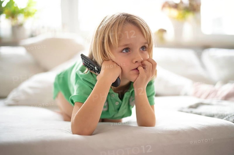 Child, boy, 7 years old, blonde, lying on sofa with remote control in hand, watching TV, watching television in front of television, excited, fascinated, Stuttgart, Baden-Württemberg, Germany