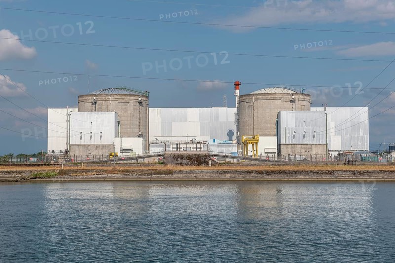 A large industrial nuclear power plant is located on a quiet water front. Two tall cylindrical structures dominate the skyline, with overhanging power lines and distant clouds that ennoble the blue sky. Fessenheim, Haut Rhin, Alsace, France