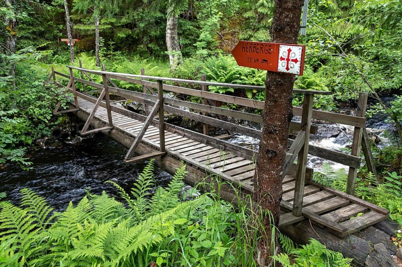 Narrow wooden bridge over a river in a green forest with a signpost to a hostel, Olav's Cross, Olav's Way or Olavsleden pilgrimage route, Norway