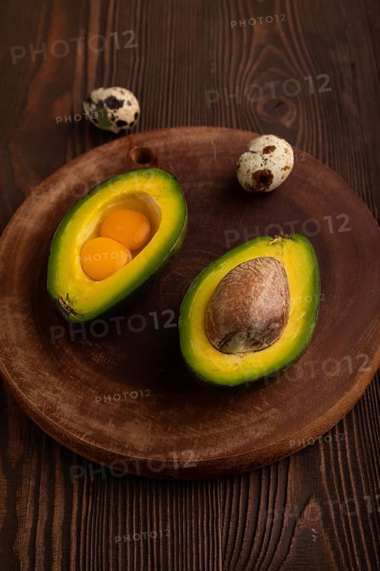 Two Avocado halves with pit and Egg yolk on round wooden cutting board on brown wooden, copy space, minimalism, low key, side view, close up