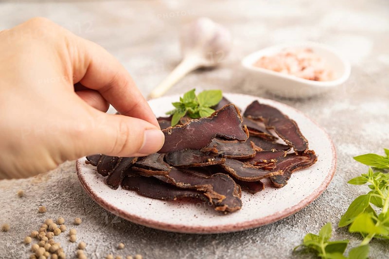 Armenian Basturma dried meat with hand on plate with pepper and herbs on brown concrete background. Side view, close up, selective focus