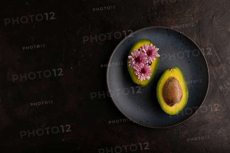 Two Avocado halves with pit and Chrysanthemums on blue plate on black concrete background, top view, flat lay, copy space, minimalism, low key