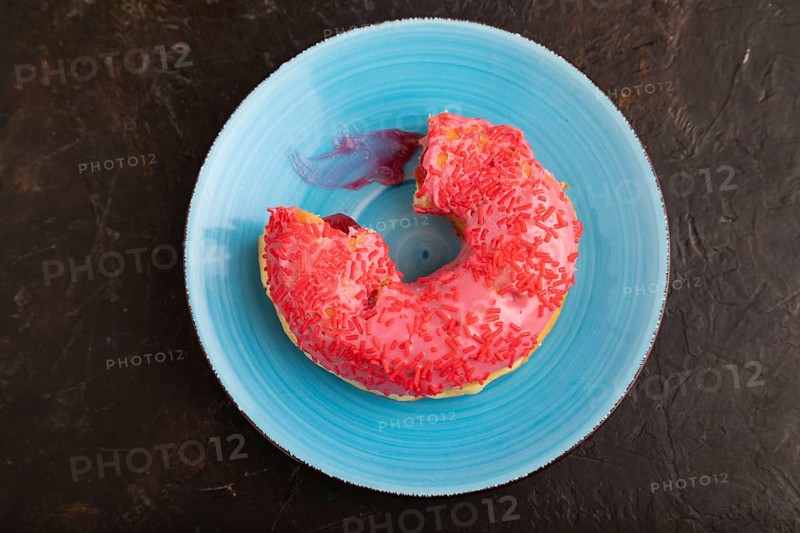 Bitten Pink Donut with sprinkles on blue ceramic plate on black concrete background, top view, flat lay, copy space, minimalism