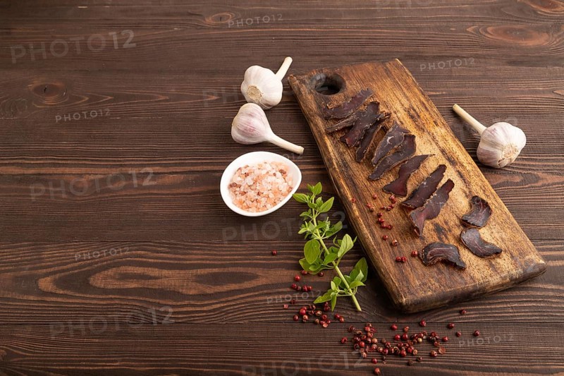 Armenian Basturma dried meat on wooden cutting board with pepper and herbs on brown wooden background. Side view, copy space