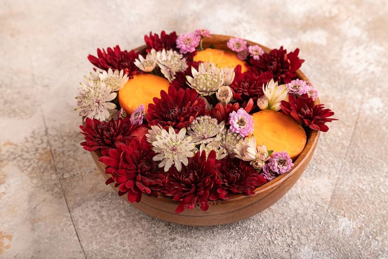 Wooden bowl with carrot slices and red Chrysanthemum flowers, Astrantia flowers, flower salad on brown concrete background, side view, close up, minimalism