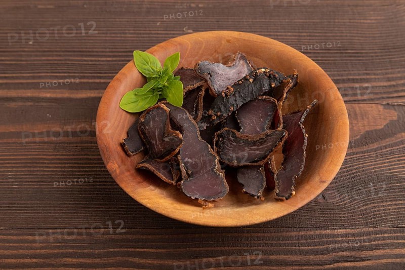 Armenian Basturma dried meat on wooden bowl with pepper and herbs on brown wooden background. Side view, copy space
