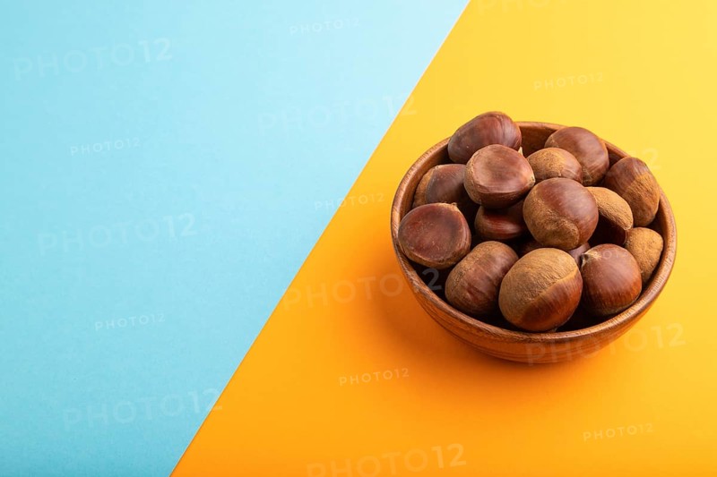 Wooden bowl with raw edible ?hestnuts on blue and orange paper pastel background, side view, copy space, minimalism