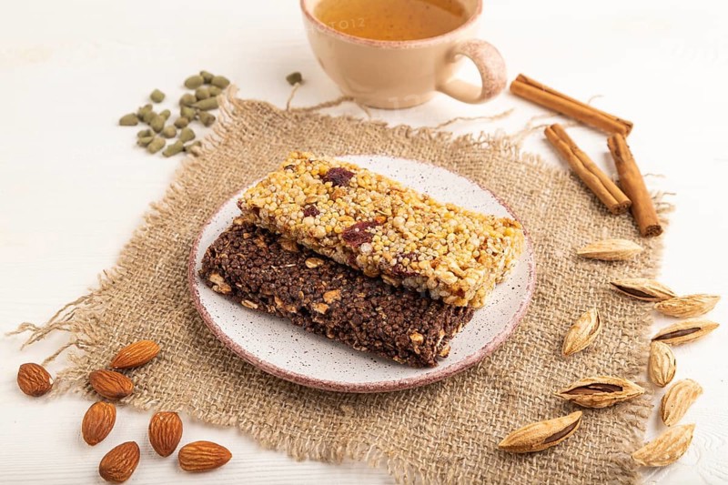 Granola bars with caramel, nuts, flakes in ceramic plate on white wooden background, beige linen napkin, cup of green tea. Side view, close up