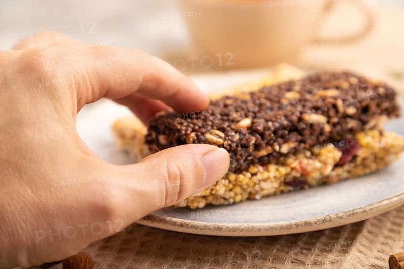 Granola bars with caramel, nuts, flakes in ceramic plate on brown concrete background with hand, beige linen napkin, cup of green tea. Side view, close up, selective focus
