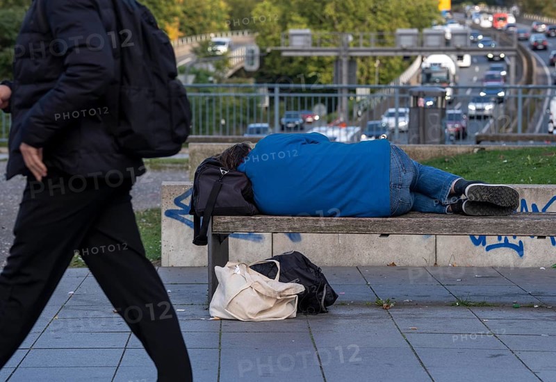 Man sleeping on a bench, travelling bag serves as pillow, in the city centre, passers-by walk by