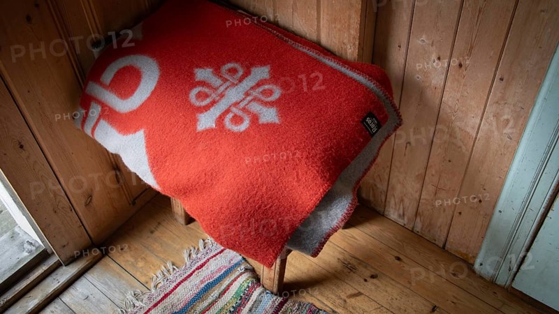 Rote Wolldecke mit Olavsweg-Symbol liegt auf einem Holzschemel, historischer Bauernhof und Pilgerunterkunft Budsjord, Dovre, Pilgerweg Olavsweg oder Olavsleden, Gudbrandsdalen oder Gudbrandstal, Norwegen
