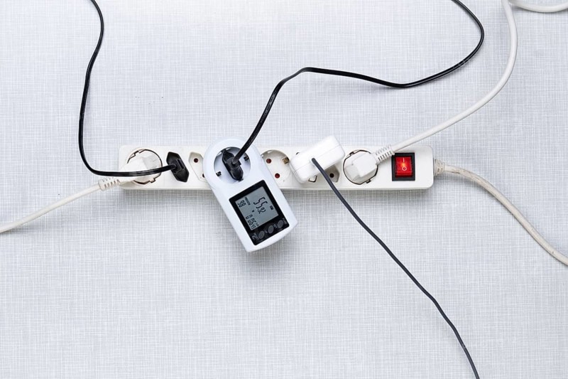 Top view of a power strip with on/off switch and various inserted plugs, cables and an electricity cost meter on a white Resopal panel