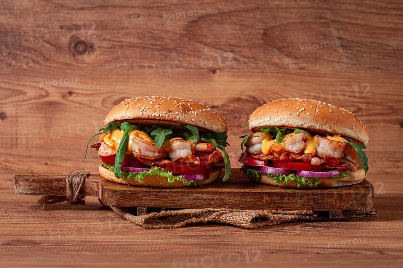 Two shrimp burgers and buns with sesame seeds, bacon, lettuce, tomatoes and onions, on a wooden board, rustic style