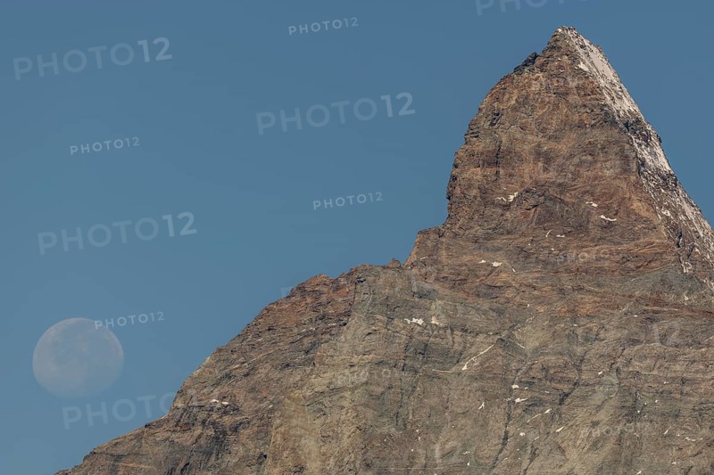 The Matterhorn mountain peak rises steeply against a bright blue sky. The full moon glistens gently above, creating a marvellous view in the serene evening hours. Matterhorn, Zermatt, Valais, Alps, Swiss
