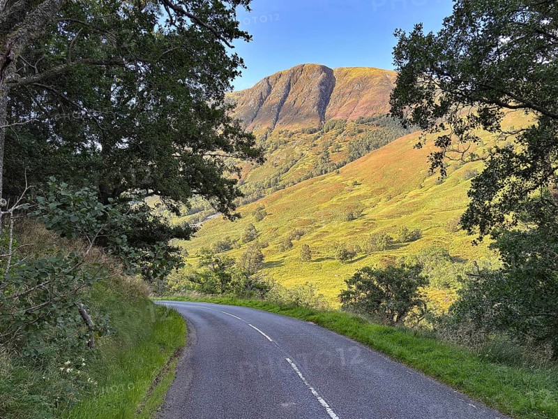 A winding road leads through a green landscape under a clear sky with a mountain in the background, Glen Nevis, Highlands, Fort William, Scotland, United Kingdom