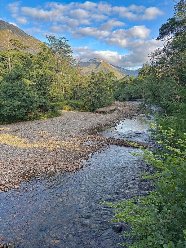 A tranquil river flows through a wooded landscape with mountains in the background and a clear blue sky, Glen Nevis, Highlands, Fort William, Scotland, United Kingdom