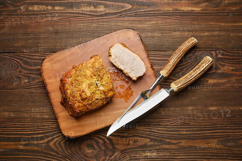 A deliciously roasted piece of pork meat on a wooden cutting board served with carving knife and fork