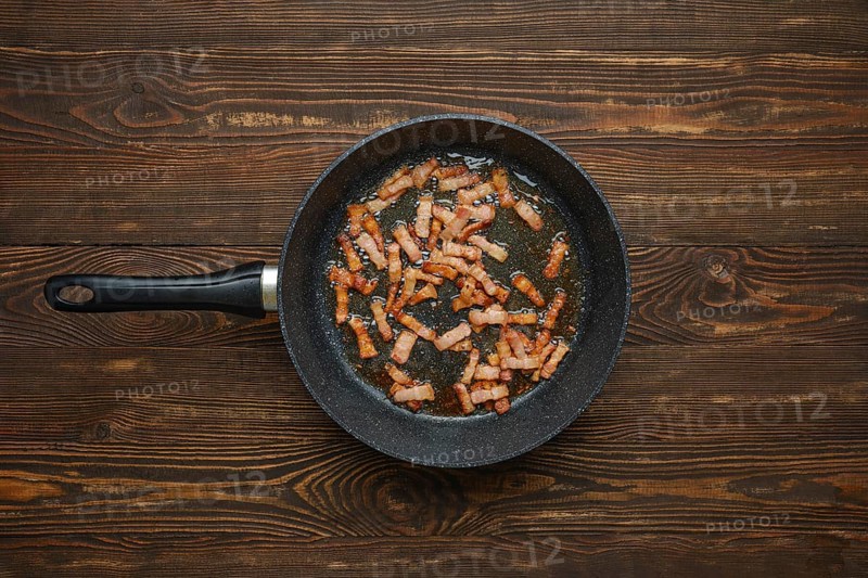 Sizzling strips of bacon fry in a black skillet, releasing savoury aroma and fat on rustic wooden tabletop