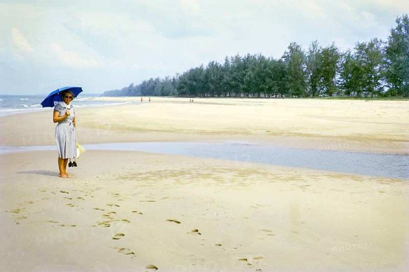 British woman holding umbrella standing on sandy beach south of estuary, Rompin, East coast, Pahang, Malaya, Malaysia, south east Asia 1964