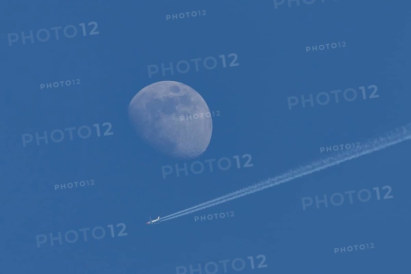Boeing 737 jet passenger aircraft of Norweign Air with a vapour trial or contrail behind flying in a blue sky passing by the moon, England, United Kingdom