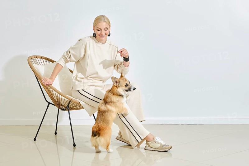 A joyful woman in a light outfit interacts with her playful corgi in a stylish indoor space. The cozy atmosphere highlights their bond, showcasing a moment of happiness and companionship