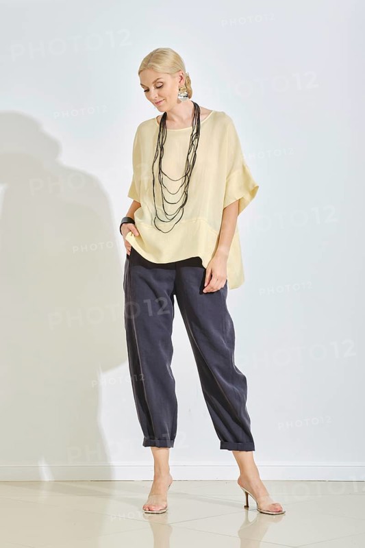 A woman stands confidently wearing a stylish yellow blouse and dark pants, accessorized with a necklace. Soft lighting highlights her relaxed pose and contemporary style