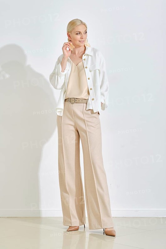 A woman dressed in a cream top and stylish beige trousers strikes a confident pose in a minimalist interior. She pairs the outfit with a light jacket, exuding elegance and sophistication
